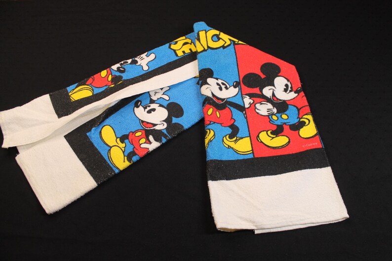 May include: A set of two white cotton towels with a colorful Mickey Mouse design. The towels feature Mickey in various poses against blue and red backgrounds, with black borders and white towel ends.