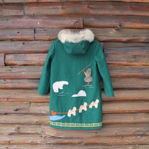 May include: A green coat with a fur-lined hood and whimsical embroidered designs. The coat features a polar bear, sled dogs, and a skier. The hem is decorated with a yellow and green pattern. The coat is hanging on a wooden background.