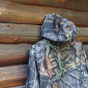 May include: A hooded camouflage jacket with a woodland pattern of green, brown, and gray tones. The jacket is hanging against a weathered wooden wall, showcasing its outdoor aesthetic. The jacket's hood is up, displaying the camouflage design.