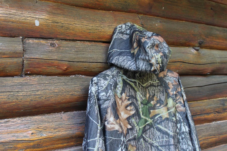 May include: A hooded camouflage jacket with a woodland pattern of green, brown, and gray tones. The jacket is hanging against a weathered wooden wall, showcasing its outdoor aesthetic. The jacket's hood is up, displaying the camouflage design.