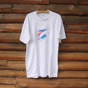 May include: White t-shirt featuring an abstract fish design in shades of blue, pink and white.
