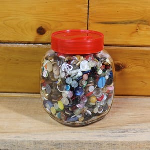 May include: A glass jar with a red lid, filled with a collection of assorted buttons. The buttons are of different sizes, shapes, and colours, including white, blue, yellow, and red. The jar is on a wooden surface.