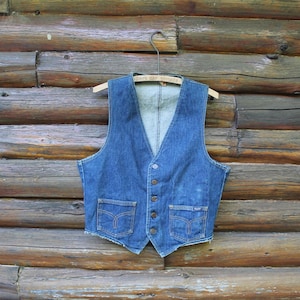 Peut inclure: Un gilet en jean bleu vintage avec un col en V, deux poches avant et des fermetures à boutons. Le gilet est suspendu à un cintre en bois sur un fond en bois rustique. Le gilet semble être en jean délavé moyen.