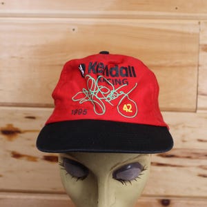 May include: Red and black baseball cap with "Kendall Racing" and "1995" printed on the front. The number "42" is also printed on the front of the cap.