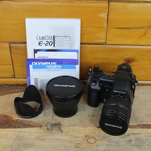 May include: A black Olympus Camedia E-20 digital camera with an attached lens, a separate lens with an Olympus cap, a black lens hood, and a white reference manual booklet with the text Camedia E-20 and Olympus printed on the cover.
