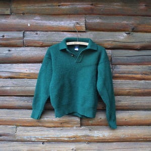 Può includere: Un maglione verde acqua con scollo a camicia e paramontura a tre bottoni. Il maglione è realizzato in un materiale a maglia spessa ed è appeso a una gruccia di legno su uno sfondo di legno rustico.