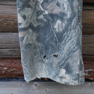 May include: A close-up view of a section of camouflage fabric, possibly denim or canvas, showing signs of wear. The fabric has a pattern of green, grey, and brown, with two holes near the bottom edge, indicating it's part of a garment.