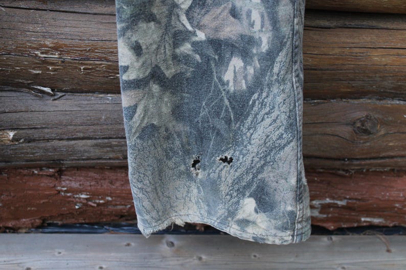 May include: A close-up view of a section of camouflage fabric, possibly denim or canvas, showing signs of wear. The fabric has a pattern of green, grey, and brown, with two holes near the bottom edge, indicating it's part of a garment.