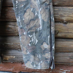 Puede incluir: Primer plano de unos pantalones de camuflaje con un estampado de hojas en tonos marr&oacute;n, gris y blanco. Los pantalones tienen una cremallera en el lateral. El fondo es una superficie de madera.