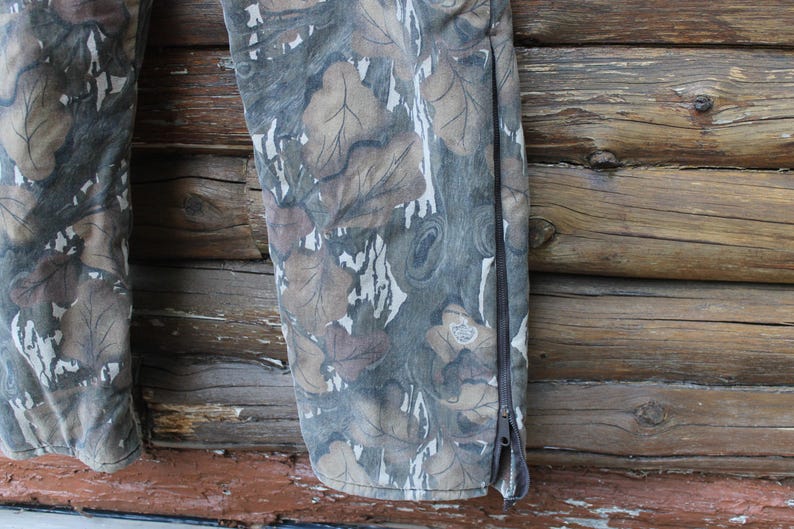 Puede incluir: Primer plano de unos pantalones de camuflaje con un estampado de hojas en tonos marr&oacute;n, gris y blanco. Los pantalones tienen una cremallera en el lateral. El fondo es una superficie de madera.