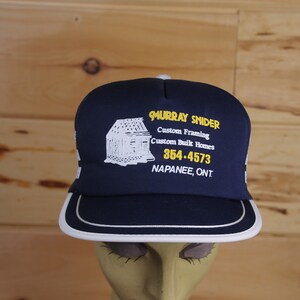 May include: A blue and white trucker hat with the text "Murray Snider Custom Framing Custom Built Homes 354.4573 Napanee, Ont" embroidered in yellow.