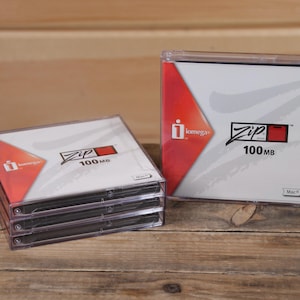May include: Several Zip 100MB diskettes in clear plastic cases. The cases have a white and red design with the "Zip" logo and "100MB" text. The image is on a wooden surface.
