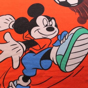 May include: Orange towel featuring Mickey Mouse kicking a soccer ball. Mickey is wearing a red shirt, blue shorts, and green and white sneakers. The soccer ball is black and white. The background is orange.