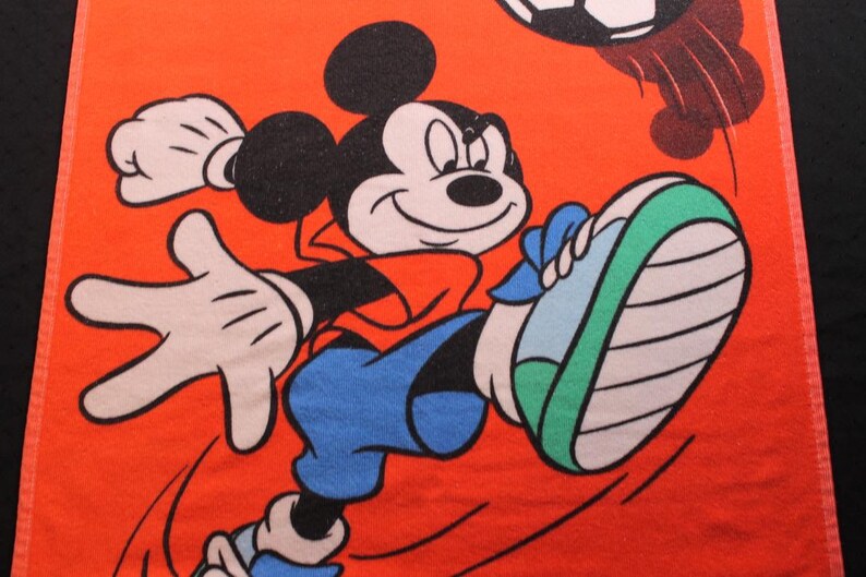 May include: Orange towel featuring Mickey Mouse kicking a soccer ball. Mickey is wearing a red shirt, blue shorts, and green and white sneakers. The soccer ball is black and white. The background is orange.