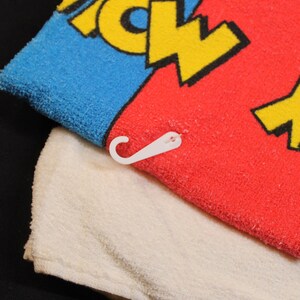 May include: A colorful towel with a blue and red section, featuring yellow text. A white towel is underneath. A white plastic hook is attached to the red towel. The towel is likely for the bathroom or beach.