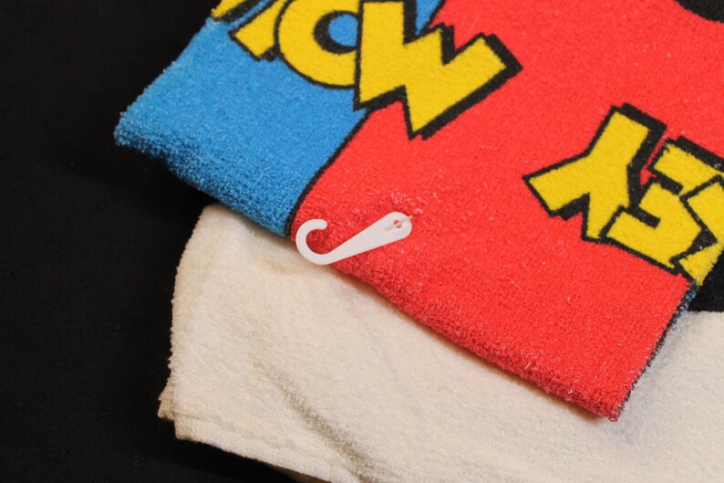 May include: A colorful towel with a blue and red section, featuring yellow text. A white towel is underneath. A white plastic hook is attached to the red towel. The towel is likely for the bathroom or beach.