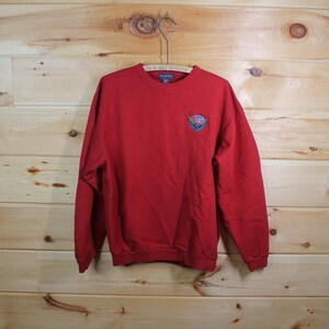 Puede incluir: Una sudadera roja de cuello redondo con un pequeño emblema bordado en el pecho izquierdo. La sudadera está colgada de una percha de metal sobre un fondo de madera. El emblema es un diseño colorido.