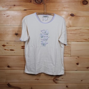 以下が含まれることがあります： 首元に紫色のトリムが付いた白いTシャツ。シャツには花柄のデザインと「Spring will come, and smiling... 」というテキストが書かれています。