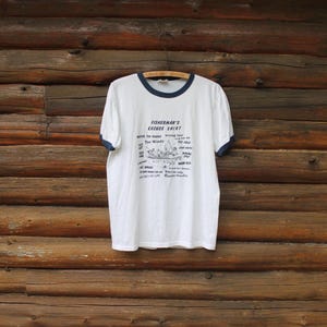 May include: A white ringer t-shirt with dark blue trim on the collar and sleeves. The front features a graphic of a boat with the text FISHERMAN'S EXCUSE SHIRT and a list of various excuses including Too Windy, Too Cold, and Wrong Bait.