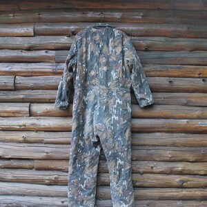 Puede incluir: Mono con estampado de camuflaje en tonos verdes, marrones y blancos, colgado frente a una pared de madera. La prenda de una pieza tiene mangas largas y un dise&ntilde;o de cuerpo entero. El patr&oacute;n de camuflaje est&aacute; dise&ntilde;ado para mezclarse con entornos forestales.