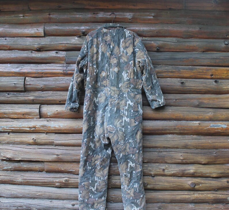 Puede incluir: Mono con estampado de camuflaje en tonos verdes, marrones y blancos, colgado frente a una pared de madera. La prenda de una pieza tiene mangas largas y un dise&ntilde;o de cuerpo entero. El patr&oacute;n de camuflaje est&aacute; dise&ntilde;ado para mezclarse con entornos forestales.
