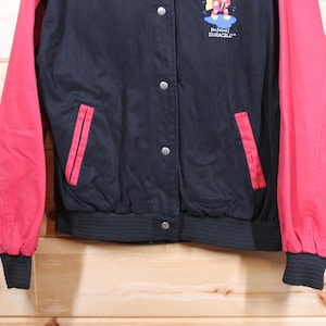K&ouml;nnte beinhalten: Eine schwarze und rote Baseballjacke mit einem Duracell-Logo und einer Comicfigur auf der Vorderseite. Die Jacke hat einen Rei&szlig;verschluss und zwei Taschen.
