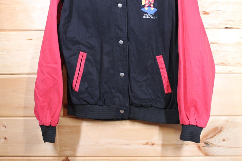 K&ouml;nnte beinhalten: Eine schwarze und rote Baseballjacke mit einem Duracell-Logo und einer Comicfigur auf der Vorderseite. Die Jacke hat einen Rei&szlig;verschluss und zwei Taschen.