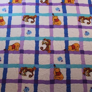 Könnte beinhalten: Ein weißer Stoff mit einem Gittermuster in Blau, Lila und Türkis. Der Stoff ist mit Cartoon-Bildern von Winnie Puuh und Tigger sowie blauen Marienkäfer-Designs verziert.