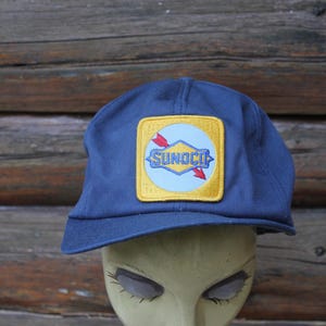Könnte beinhalten: Marineblaue Baseballkappe mit einem quadratischen Aufnäher mit dem Sunoco-Logo. Der Aufnäher hat einen gelben Rand, einen hellblauen Kreis und den Namen Sunoco in Weiß. Rote Pfeile zeigen zur Mitte.