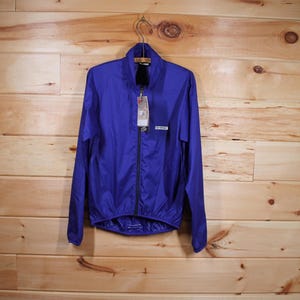 Puede incluir: Una chaqueta ligera de color morado con cierre de cremallera y un pequeño logotipo en el pecho izquierdo. La chaqueta tiene un cuello alto y mangas largas.