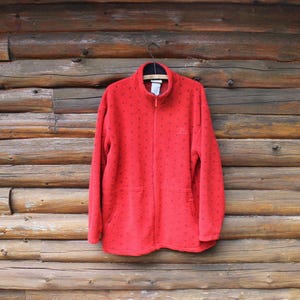 May include: A bright red fleece jacket featuring a full-length front zipper, a high collar, and a repeating pattern of small dark blue star shapes. The jacket includes long sleeves and two front pockets, with a small embroidered logo on the chest.
