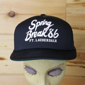 May include: Black trucker hat with the text "Spring Break '86 Ft. Lauderdale" in white script. The hat has a black brim and mesh back. The hat is displayed on a mannequin head.