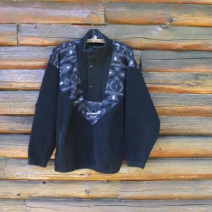 Puede incluir: Sudadera de vellón negra con un patrón geométrico en la parte delantera. La sudadera tiene un cierre de botones a presión en el cuello y un bolsillo en el lado izquierdo. El nombre de la marca "Goodwear" es visible en la parte delantera.