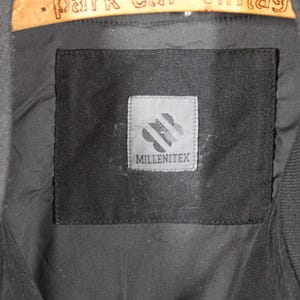 K&ouml;nnte beinhalten: Eine schwarze Jacke mit einem schwarzen Etikett, auf dem "Millenitex" steht, und einem wei&szlig;en Logo mit einem stilisierten Blumendesign.