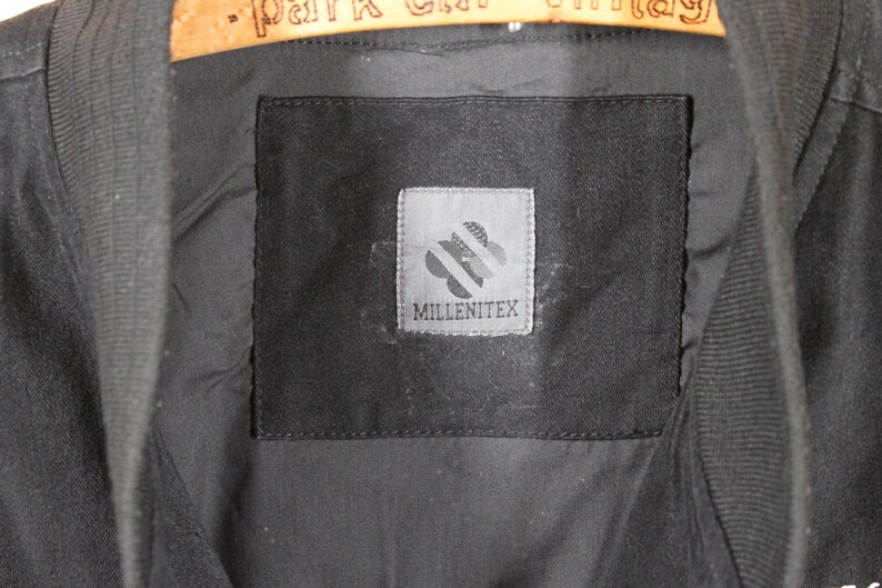 K&ouml;nnte beinhalten: Eine schwarze Jacke mit einem schwarzen Etikett, auf dem "Millenitex" steht, und einem wei&szlig;en Logo mit einem stilisierten Blumendesign.