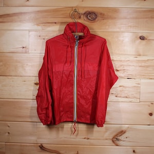 Op de afbeelding: Een rode nylon windjack met een ritssluiting en een wit, zwart en geel streepdetail op de rits. De jas heeft een capuchon en een trekkoord in de taille.