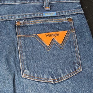 May include: A pair of blue denim jeans with a back pocket featuring a brown leather patch with the word "Wrangler" in white lettering.