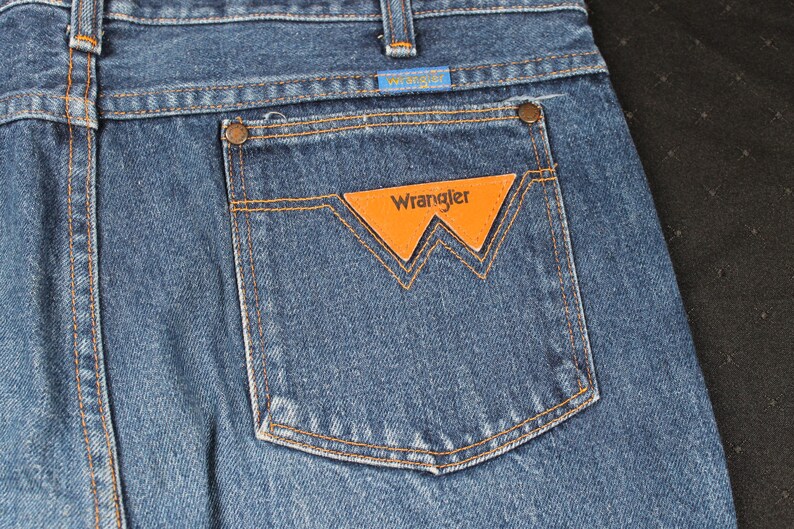 May include: A pair of blue denim jeans with a back pocket featuring a brown leather patch with the word "Wrangler" in white lettering.