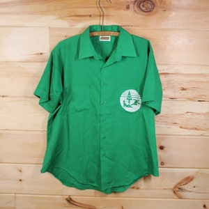 Può includere: Una camicia verde a maniche corte con bottoni e un logo bianco con un albero e persone. Il logo si trova sul lato sinistro della camicia.