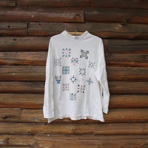 May include: A white long-sleeved top with a mock neck, featuring a pattern of floral and geometric designs in blue, pink, and green. The garment is displayed against a backdrop of rustic wooden logs.