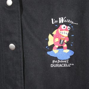 K&ouml;nnte beinhalten: Schwarze Jacke mit einem roten und blauen gestickten Design eines Cartoon-Fischs, der eine Duracell-Batterie h&auml;lt. Der Text "I'm Walkin... Yes Indeed! DURACELL" ist ebenfalls auf die Jacke gestickt.