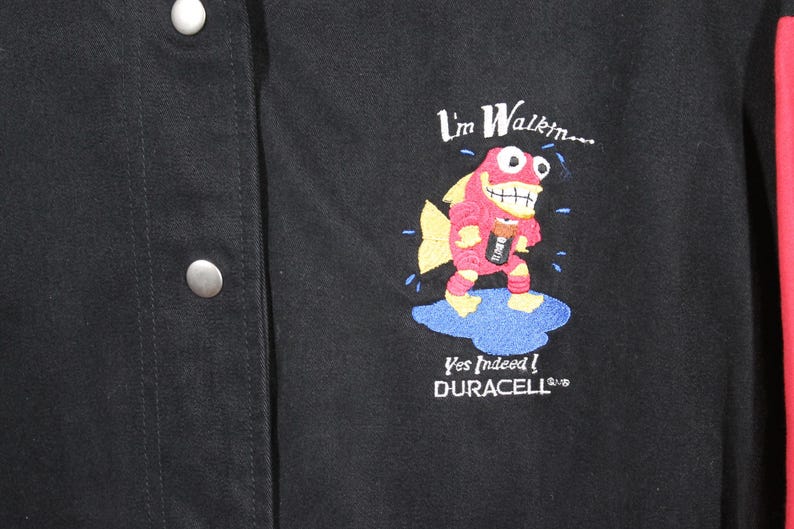 K&ouml;nnte beinhalten: Schwarze Jacke mit einem roten und blauen gestickten Design eines Cartoon-Fischs, der eine Duracell-Batterie h&auml;lt. Der Text "I'm Walkin... Yes Indeed! DURACELL" ist ebenfalls auf die Jacke gestickt.