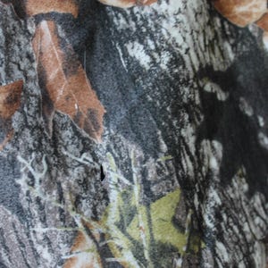 May include: Close-up of a camouflage pattern fabric. The design features a mix of brown, green, gray, and black, resembling leaves and branches. The fabric appears to be textured, with visible details in the print.