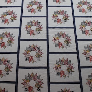 May include: A quilt featuring a repeating pattern of white square blocks separated by dark blue sashing. Each white block contains a multi-colored star motif with a circular center, composed of various floral and geometric fabric prints in red, blue, and green.