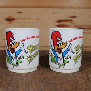 Op de afbeelding: Twee witte keramische mokken met een cartoon afbeelding van Woody Woodpecker. De mokken hebben de tekst "Ha-Ha-Ha-Ha" en "Woody Woodpecker" erop gedrukt.