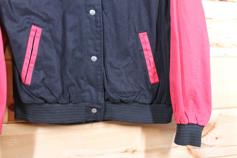 K&ouml;nnte beinhalten: Eine schwarze und rote Collegejacke mit Rei&szlig;verschluss und zwei Taschen. Die Jacke hat einen gerippten Strickkragen, B&uuml;ndchen und einen Bund.