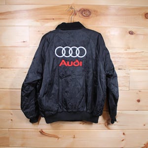 May include: Black satin bomber jacket with an embroidered Audi logo on the back. The logo features four interlocking rings in white and the word "Audi" in red.
