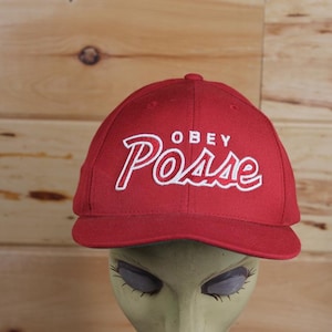 Peut inclure: Casquette de baseball rouge avec un logo brodé blanc qui dit "OBEY Posse".