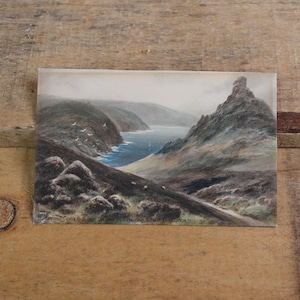May include: A vintage landscape painting depicts a coastal scene with rolling hills, a winding road, and a body of water. The artwork features muted tones of green, gray, and blue, with a flock of birds in the sky. The painting is on a rectangular piece of paper.