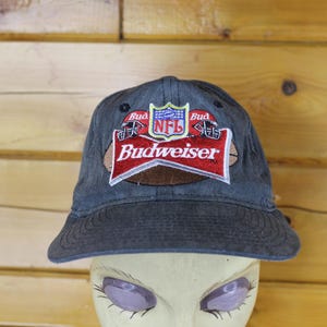 May include: A dark grey baseball cap featuring an embroidered patch on the front. The patch displays the Budweiser logo in red and white, an NFL shield logo, and two football helmets. The cap has a curved brim and a structured crown design.